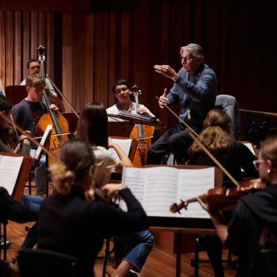 A man with grey hair and a blue jumper conducts an orchestra
